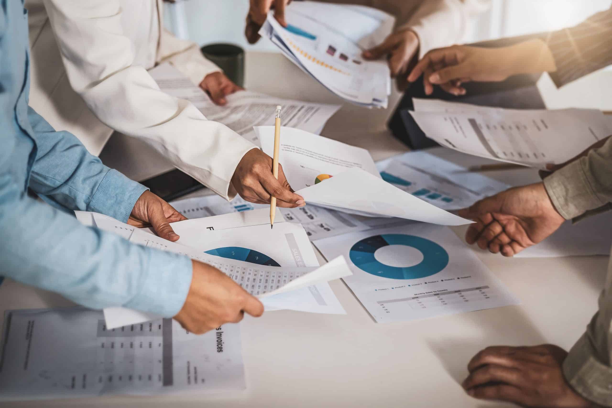 Mehrere Personen versammeln sich um einen Tisch, um gedruckte Tabellen, Diagramme und Finanzdokumente zu prüfen und zu besprechen, wobei sie mit den Händen auf etwas zeigen und Papiere weiterreichen.