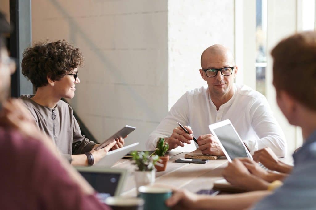 Vier Personen sitzen um einen Tisch in einem hellen Büro und diskutieren. Sie haben digitale Geräte, Notebooks und Kaffeetassen vor sich stehen. Ein Mann in Weiß scheint das Gespräch zu leiten.