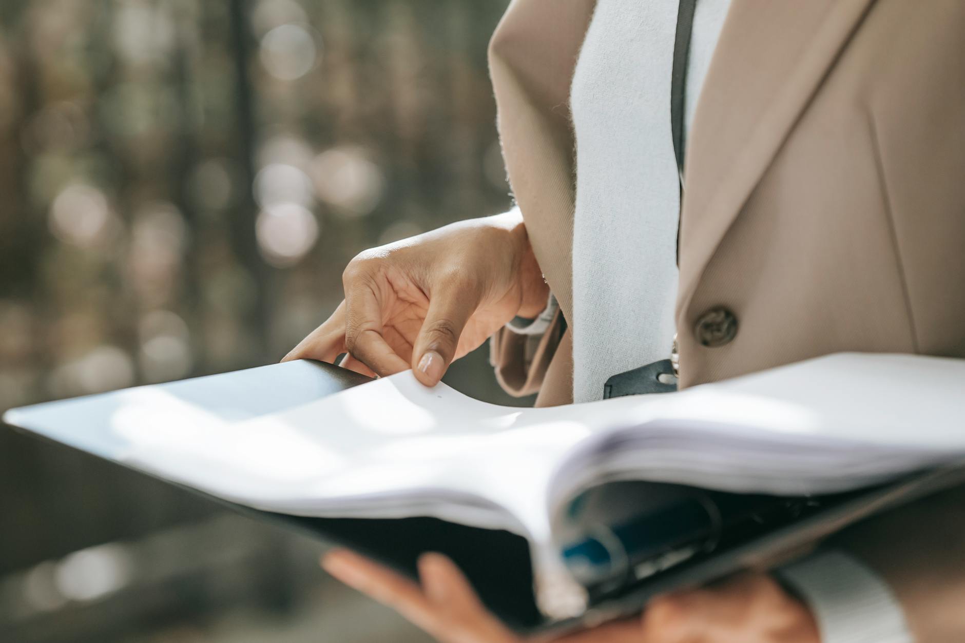 Eine Person, die einen beigen Blazer trägt, hält einen großen aufgeschlagenen Ordner oder eine Mappe in der Hand und blättert darin, als würde sie Dokumente oder Papierkram durchsehen.