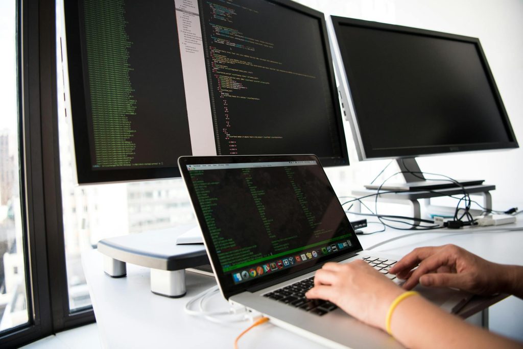 In einem hellen Büro mit großen Fenstern tippt eine Person auf einem Laptop, der an zwei große Monitore angeschlossen ist, auf denen Codezeilen angezeigt werden.