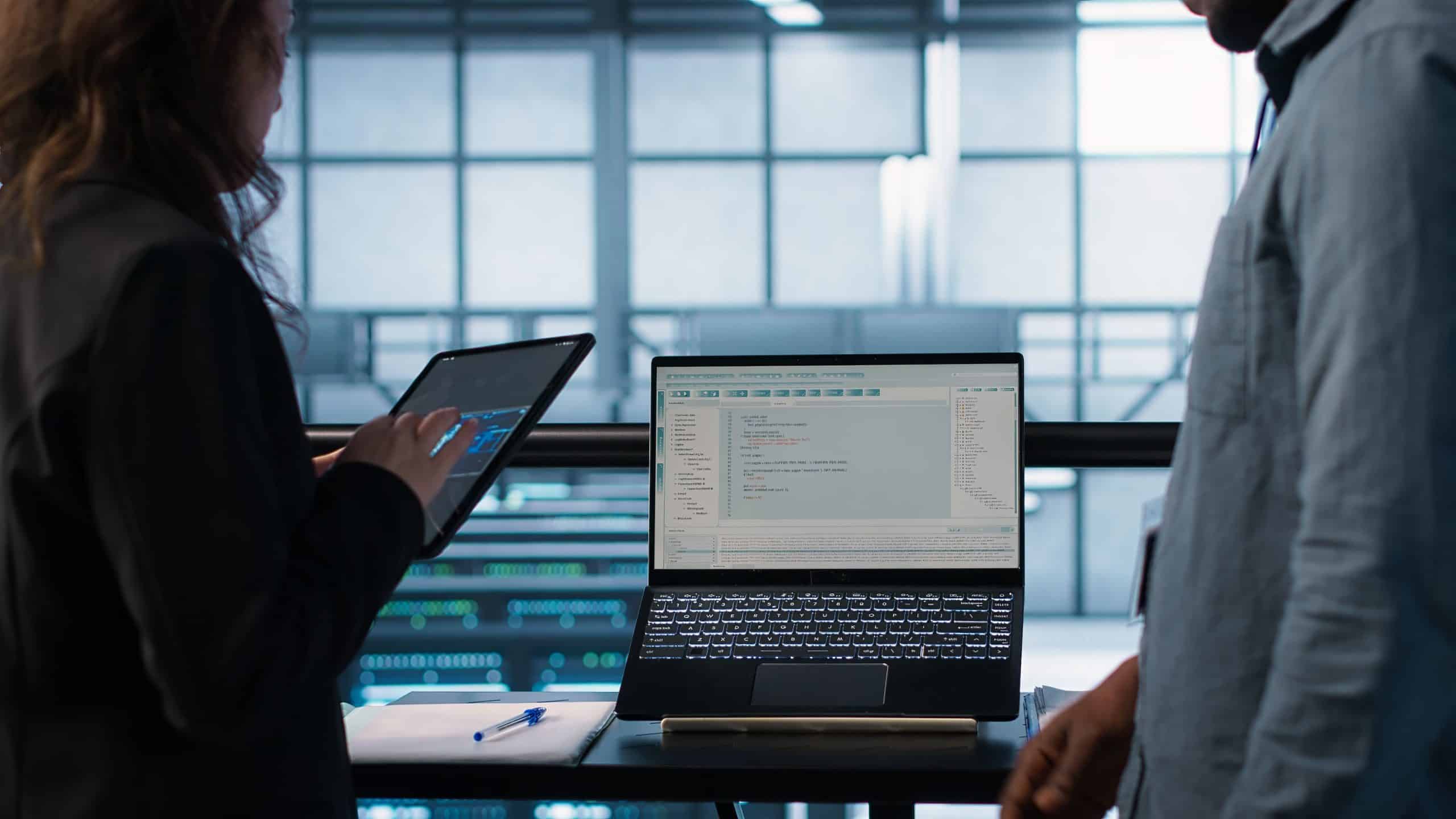 Zwei Personen stehen an einem Tisch mit einem Laptop, auf dessen Bildschirm ein Code angezeigt wird. Eine Person hält ein Tablet. Die Umgebung scheint ein modernes Büro oder Rechenzentrum mit großen Fenstern und sichtbaren Geräten zu sein.