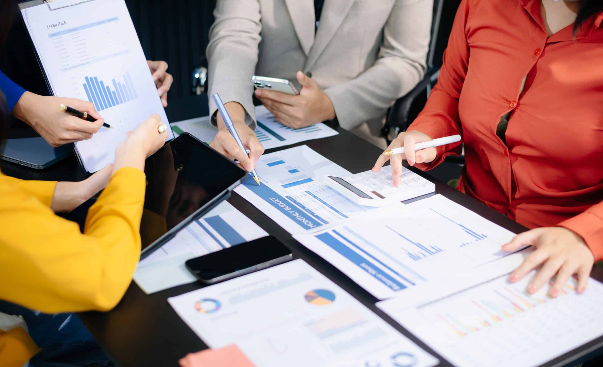 Vier Personen in Geschäftskleidung sitzen um einen Tisch, der mit Diagrammen, Schaubildern und Dokumenten bedeckt ist, und diskutieren Daten und machen sich Notizen mit Stiften und Tablets.