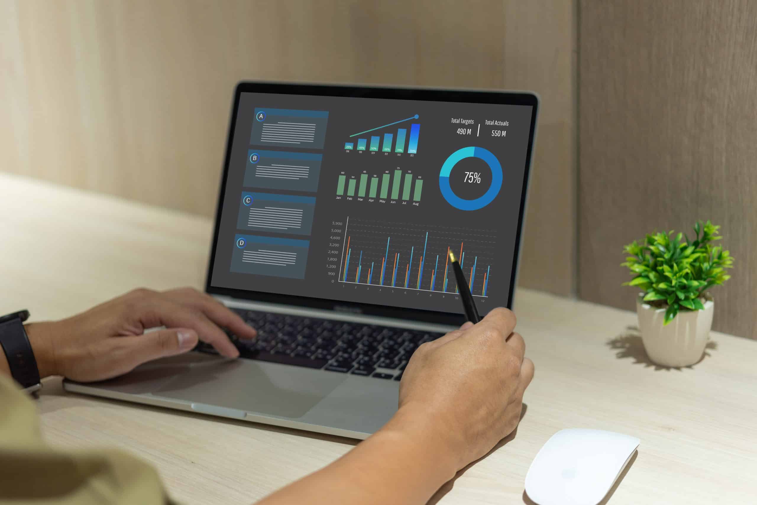 Eine Person benutzt einen Laptop, auf dem Datenanalysegrafiken und Diagramme angezeigt werden, und zeigt mit einem Stift auf den Bildschirm. Die Szene enthält eine Maus und eine kleine Topfpflanze auf einem hellen Holztisch.