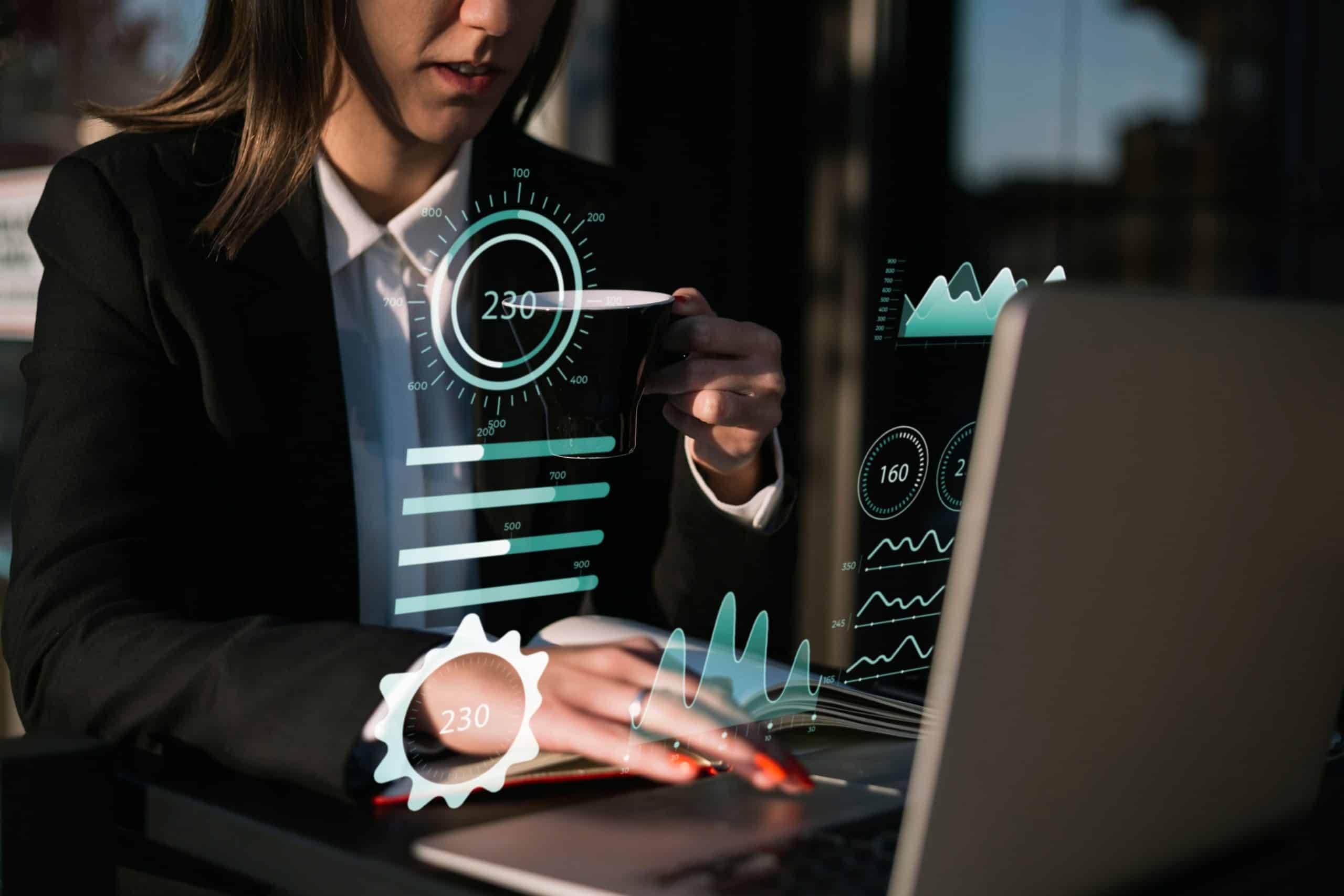 Eine Frau in Geschäftskleidung arbeitet an einem Tisch an einem Laptop und hält eine Tasse in der Hand. Vor ihr werden futuristische digitale Diagramme und Datenvisualisierungen projiziert, die auf Datenanalyse oder Technologieeinsatz hinweisen.