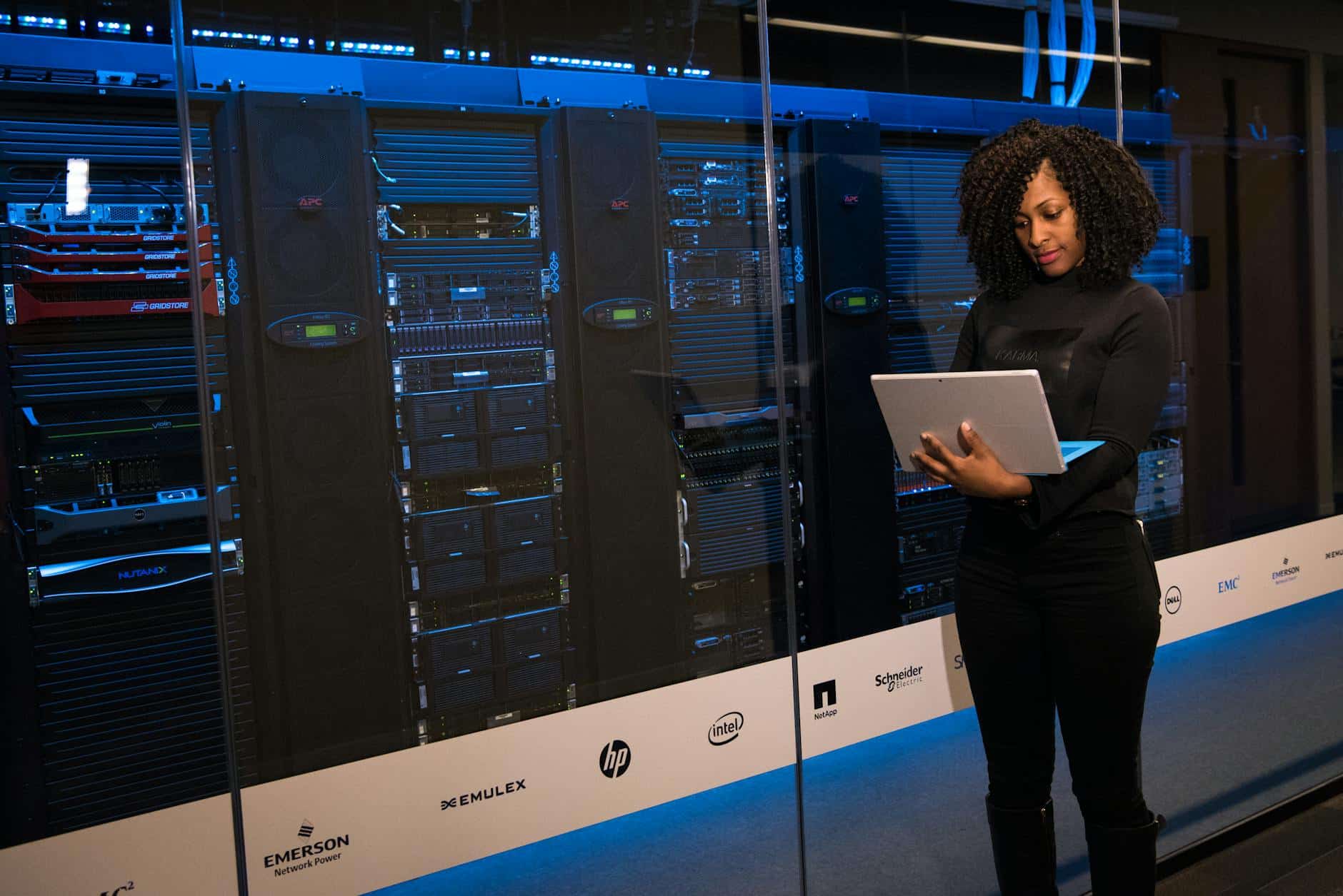 Eine Frau steht vor einem Server-Rack in einem Rechenzentrum und arbeitet an einem Laptop. Sie trägt ein schwarzes Outfit und ist auf den Bildschirm konzentriert. Im Serverraum sind verschiedene Geräte und Firmenlogos ausgestellt.
